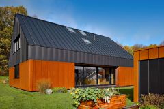 Kompaktní fasáda jednoduchého domu je členěna vertikálními falcovanými spoji cortenového a černého plechu. Barevný kontrast doplňují prosklené plochy